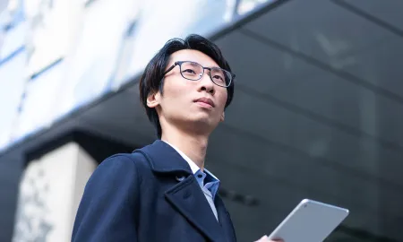 A man with glasses in a dark coat looking thoughtfully upward while holding a tablet in front of a modern building.