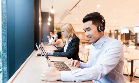 A male business student wearing a headset and working on a laptop in a modern campus study lounge.