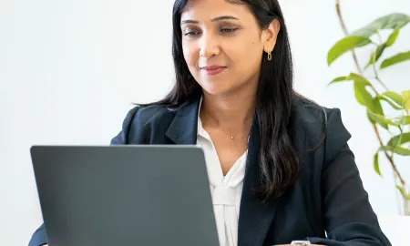 A business student in a blazer focused on her laptop while studying in a bright, plant-filled workspace.