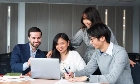 A diverse group of four business school students smiling and collaborating around a laptop.