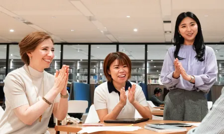 Three female business school students smiling and clapping around a table in a study area.
