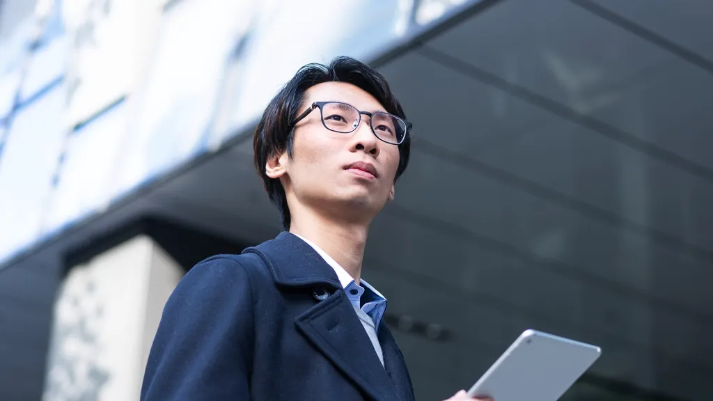 A man with glasses in a dark coat looking thoughtfully upward while holding a tablet in front of a modern building.