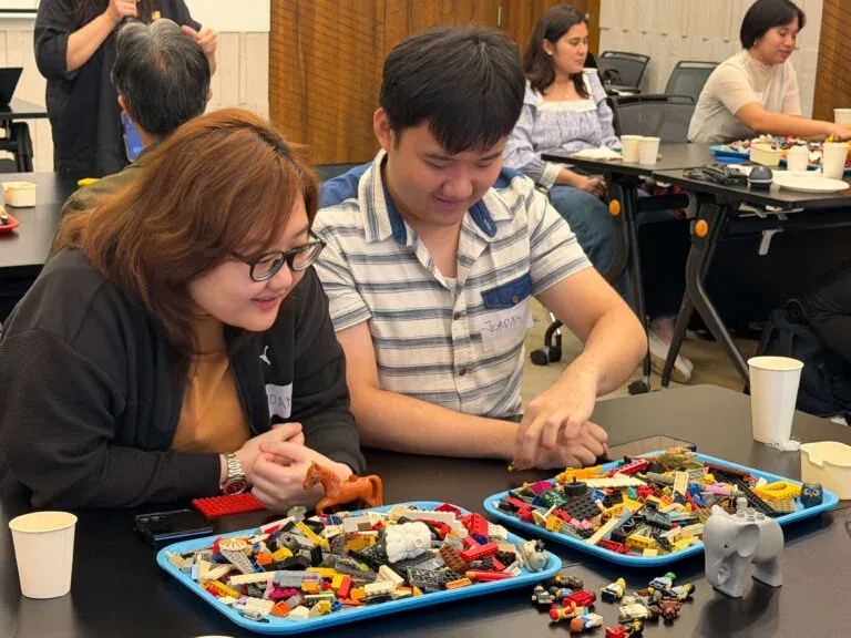 Two participants building from trays of loose bricks at their table.