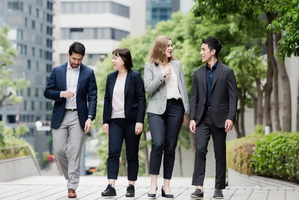 Four diverse business students in professional attire walking and conversing outdoors on a city campus.