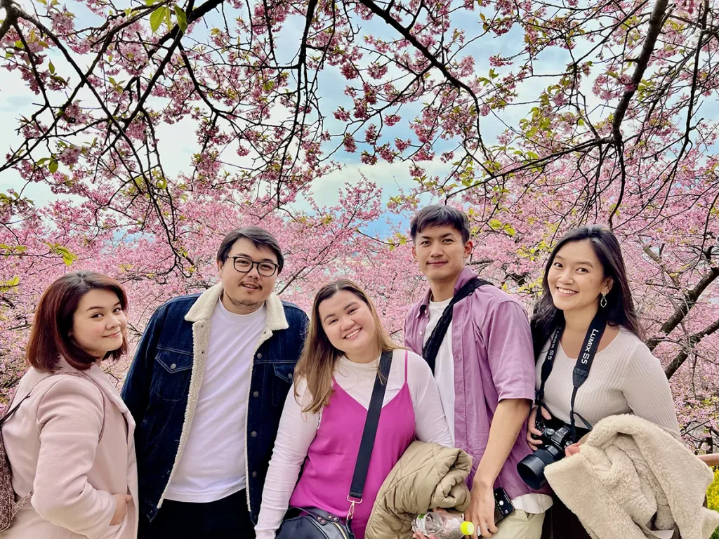 Five people smiling and posing together outdoors under blooming pink cherry blossom trees.
