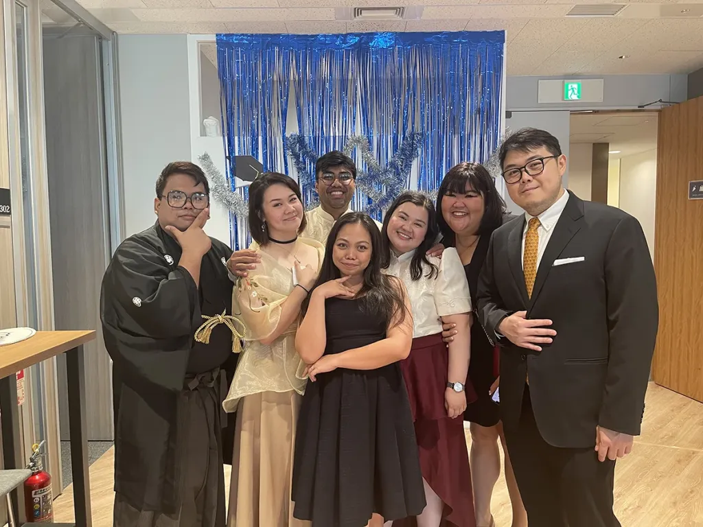 Seven business school students in formal and traditional attire smiling together in front of a blue tinsel photo backdrop.