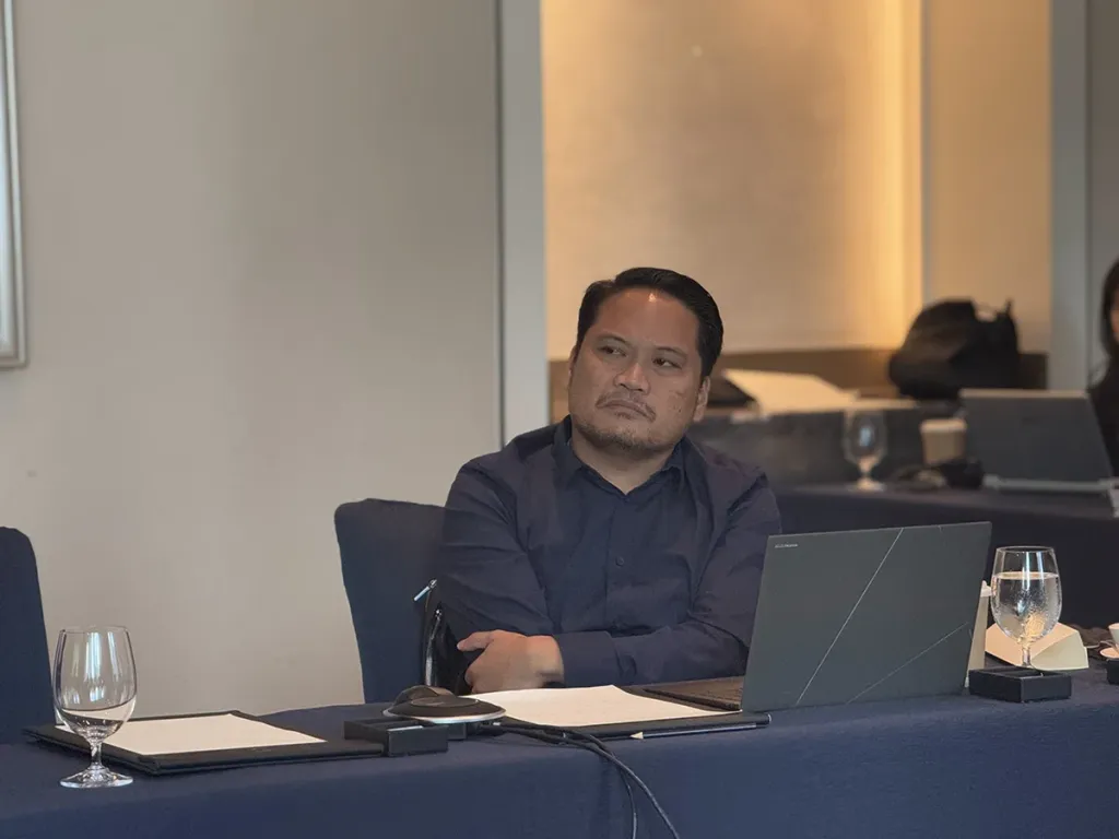 A male student sitting at a table with an open laptop, listening attentively during a business school class.