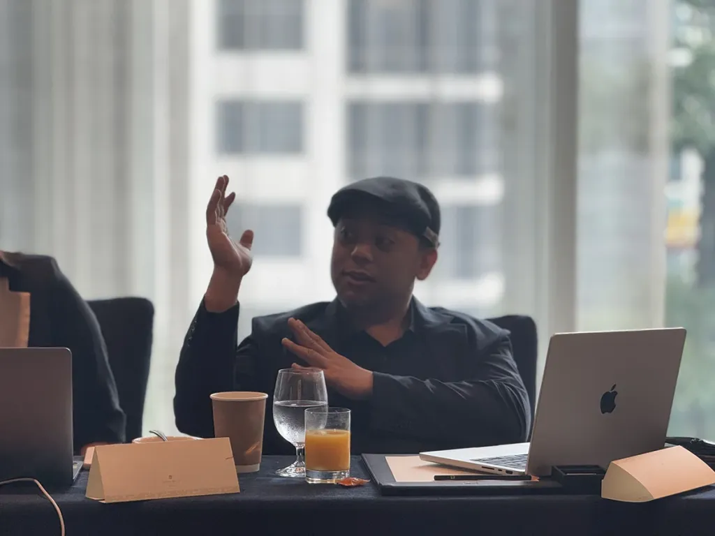 A male business school student wearing a flat cap gestures while speaking at a conference table with an open laptop.