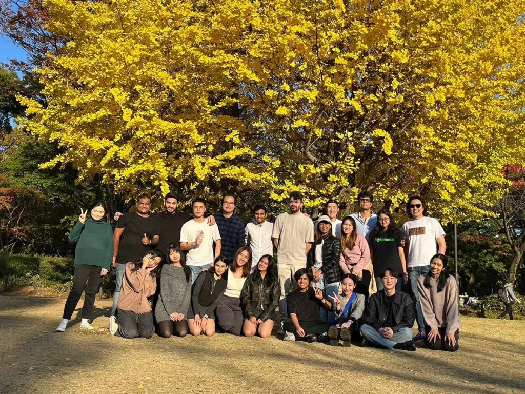 A diverse group of business school students posing together outdoors in front of a large, vibrant yellow ginkgo tree.