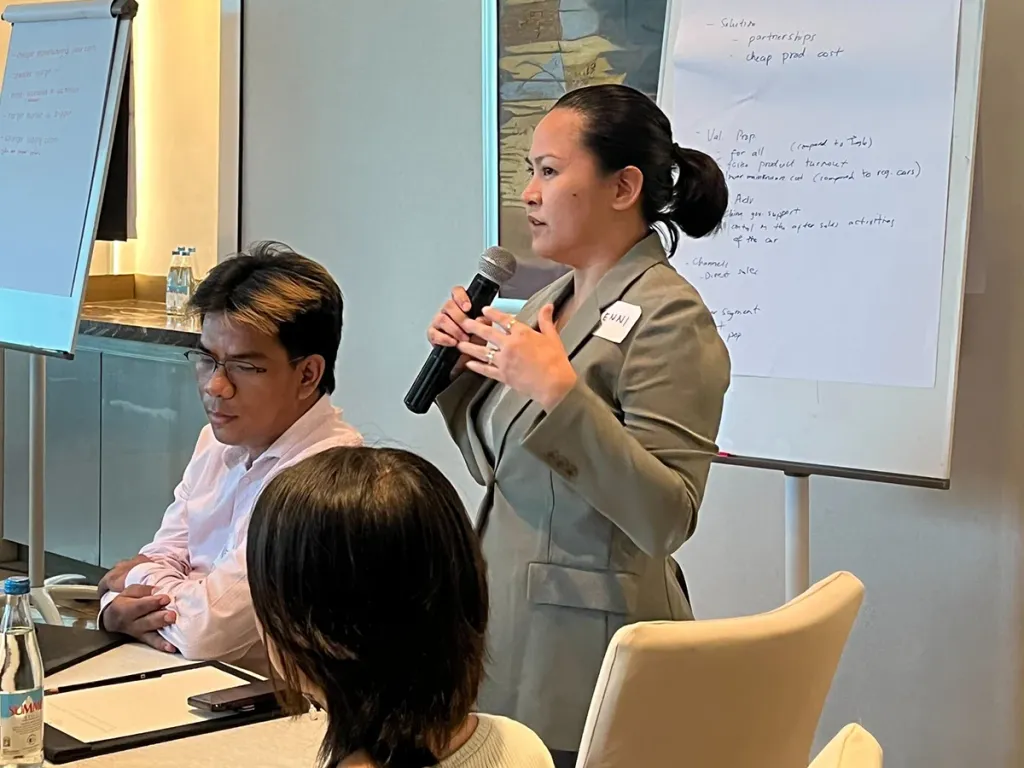 A female student standing and speaking into a microphone during a Pre-MBA class, with a written flipchart behind her.