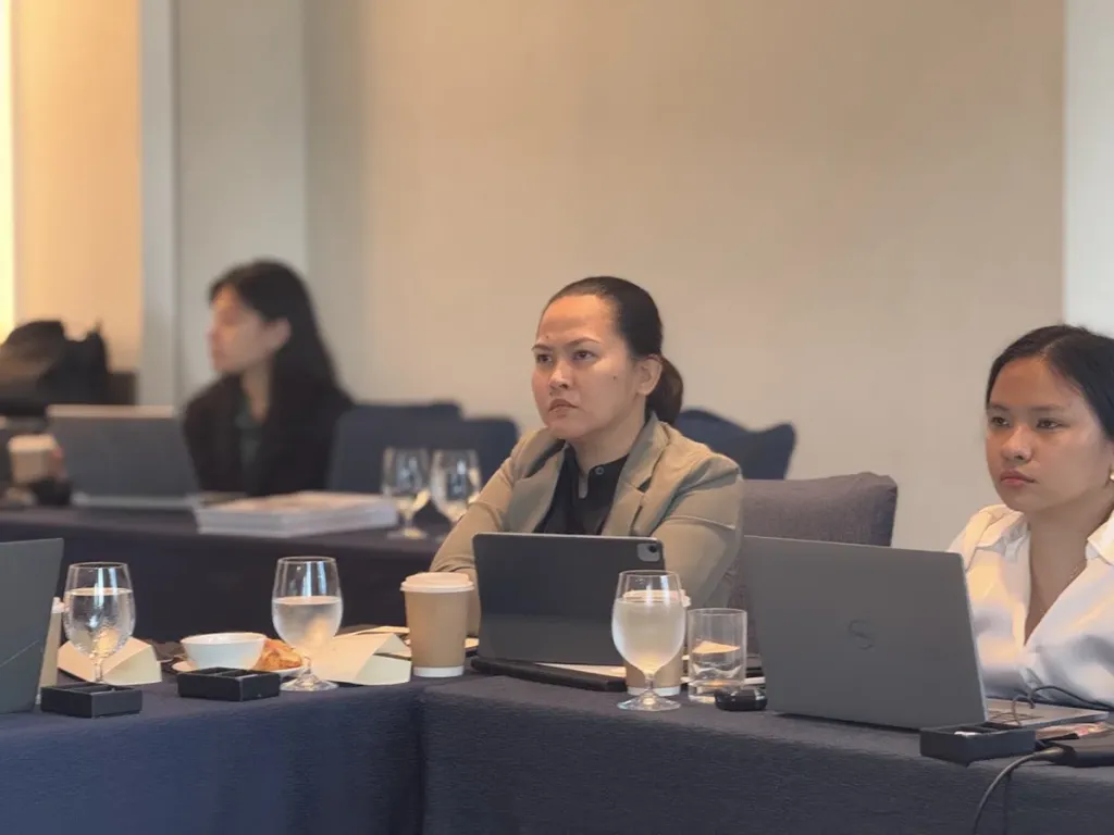 Three students sitting at a conference table with laptops and beverages, listening attentively during a Pre-MBA class.