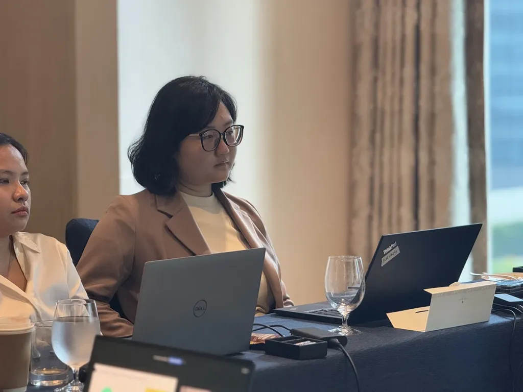 A female student with glasses sits at a table with laptops, listening attentively during a Pre-MBA class.