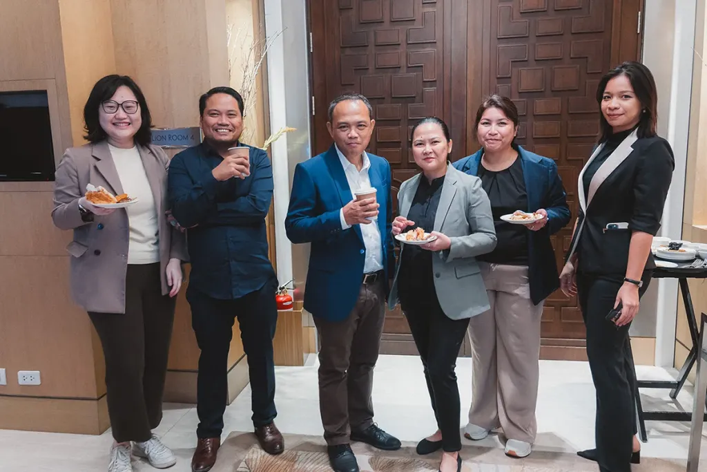 Six Pre-MBA students stand smiling and posing together with coffee and snacks during a networking break.