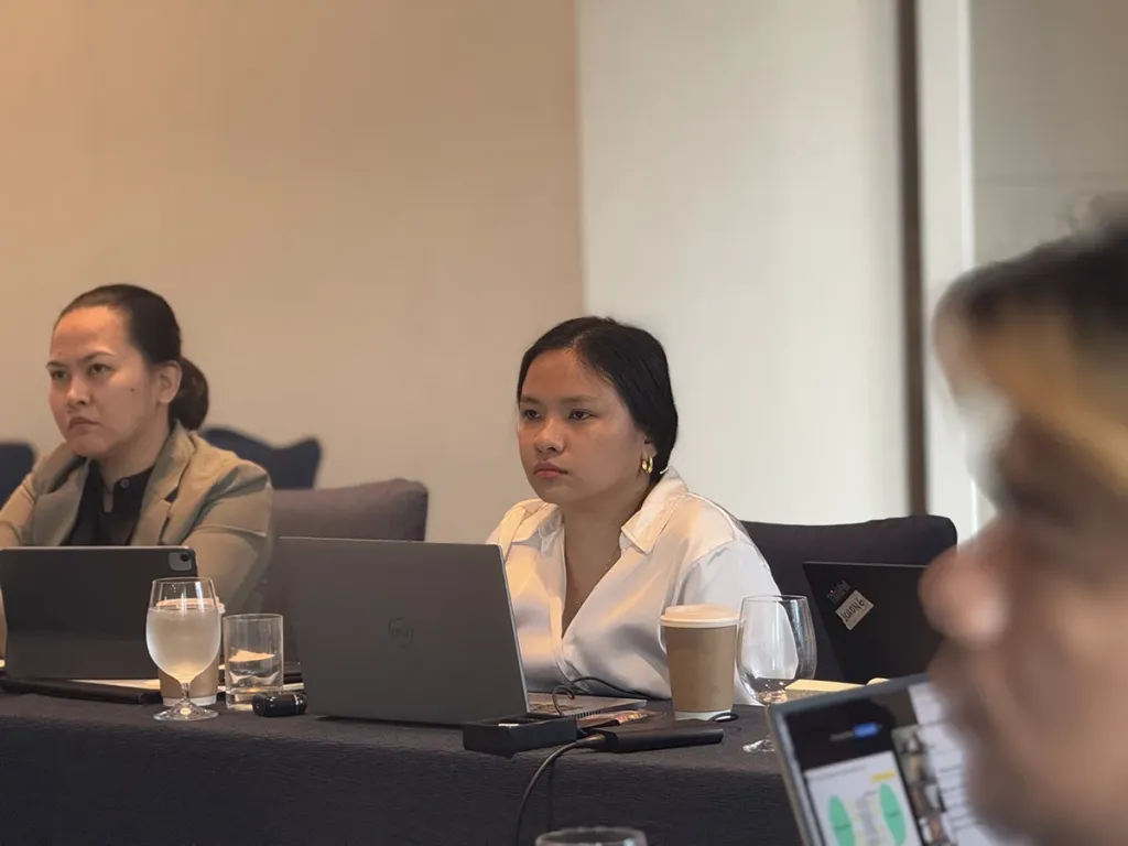 A female student in a white blouse sitting at a conference table with an open laptop, listening attentively during a class.