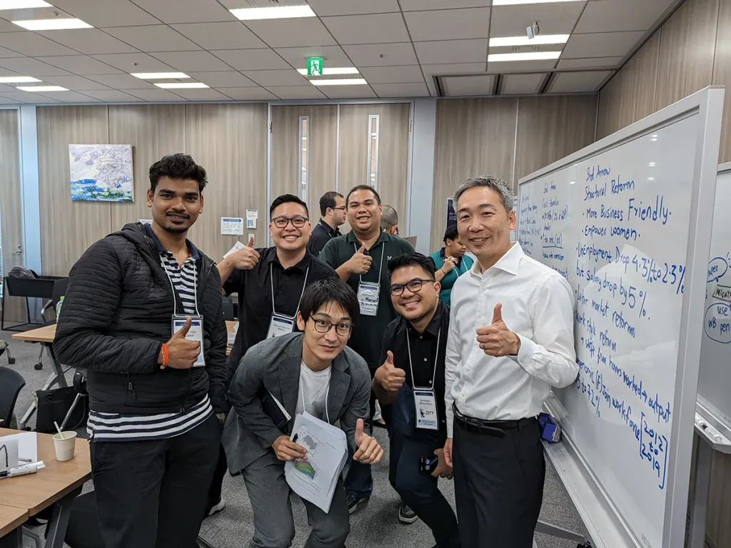 Five smiling men and their lecturer pose with thumbs up next to a whiteboard in a classroom.