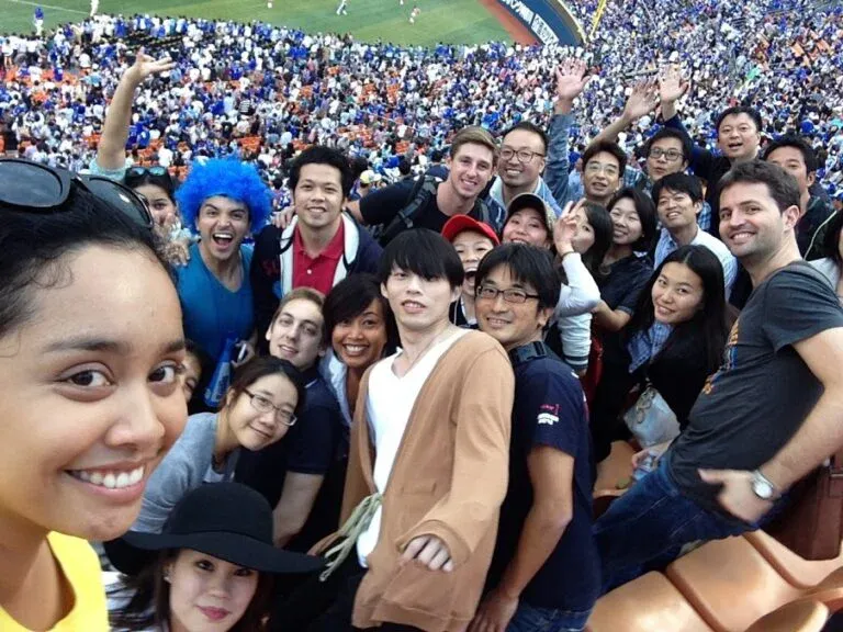 A large group of business school students taking a smiling selfie together in the stands of a crowded stadium.